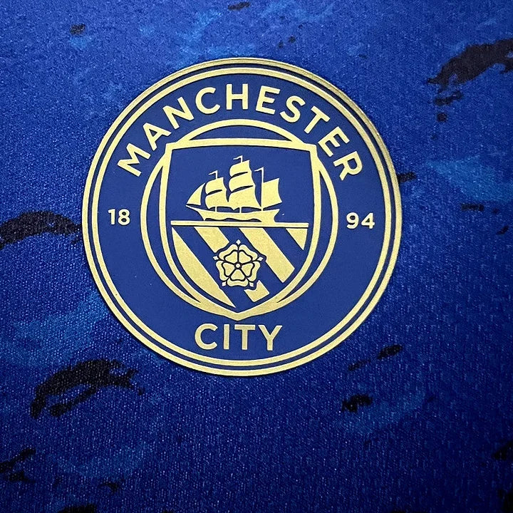 Maillot Manchester City 2023/24 Le 12 -ème Maillot, maillot de football bleu avec logo du club et détails en blanc.